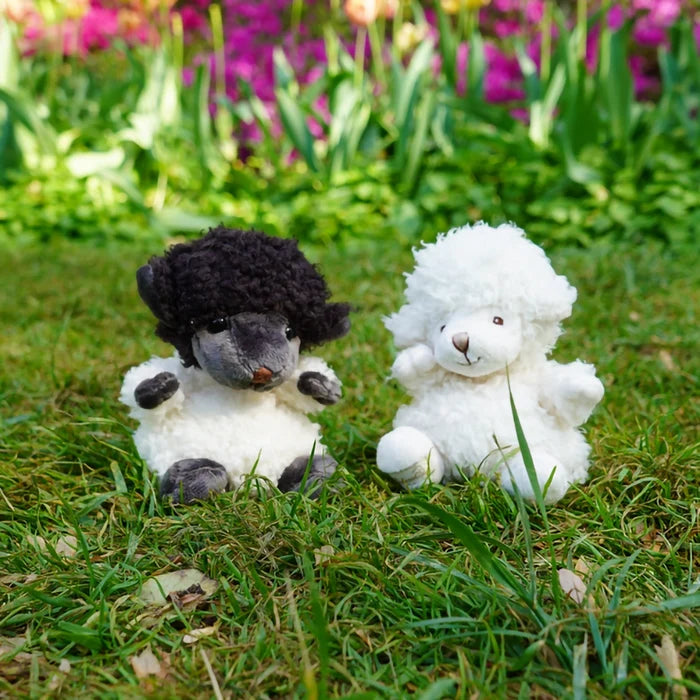 Bukowski Black Sheep plush sitting on grass next to white sheep plush in outdoor garden setting
