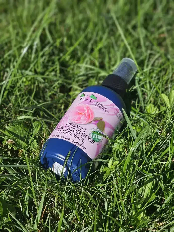 A bottle of BioMark Organic Amasculus Rose Hydrosol massage oil lies on green grass. The bottle is blue with a pink label featuring a rose graphic and a black spray nozzle.