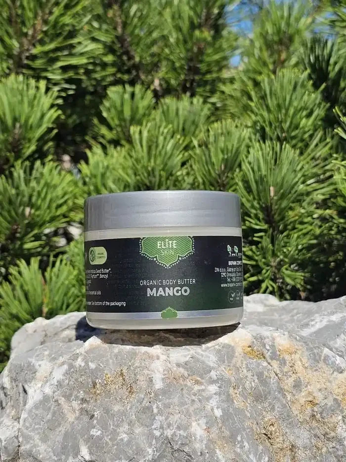 A jar of Elite Skin Organic Body Butter in Mango, placed on a gray stone with a background of green pine trees.