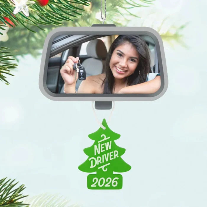 Car mirror with a reflection of a woman holding keys, Christmas tree ornament with 'New Driver 2026' text, and pine branches.