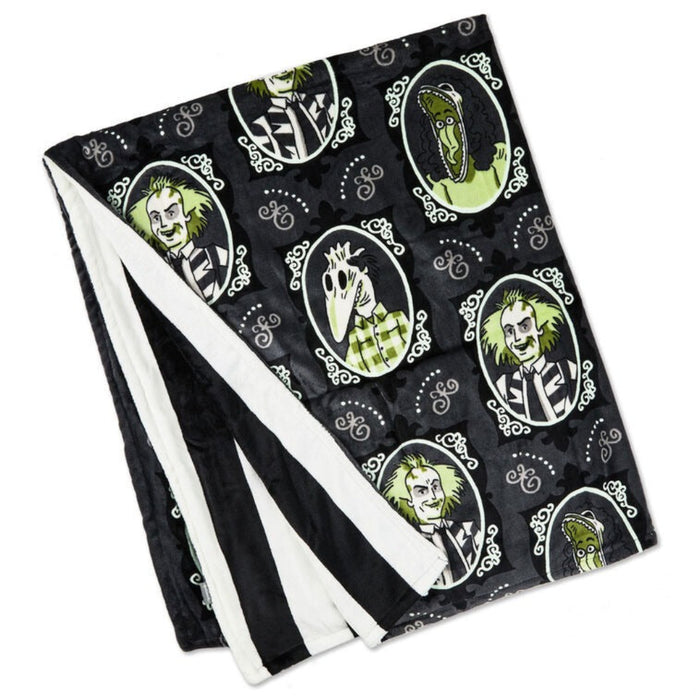 Glow-in-the-dark Beetlejuice fleece blanket featuring black and gray framed portraits of Beetlejuice, Lydia, Delia, Barbara, and Adam, with green glowing accents and a black-and-white striped back.