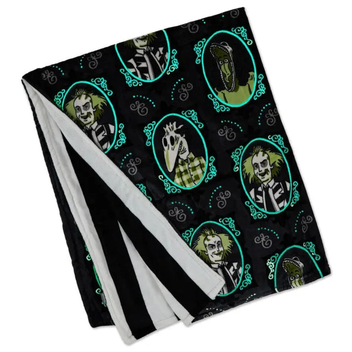 Glow-in-the-dark Beetlejuice fleece blanket featuring black and gray framed portraits of Beetlejuice, Lydia, Delia, Barbara, and Adam, with green glowing accents and a black-and-white striped back.