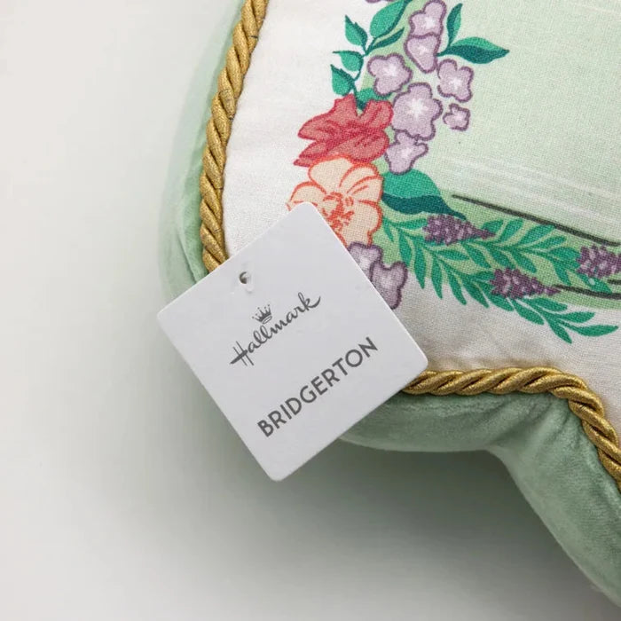 Close-up of a Bridgerton decorative pillow corner showing floral fabric, gold rope trim, and a hanging Hallmark Bridgerton product tag on a white background.