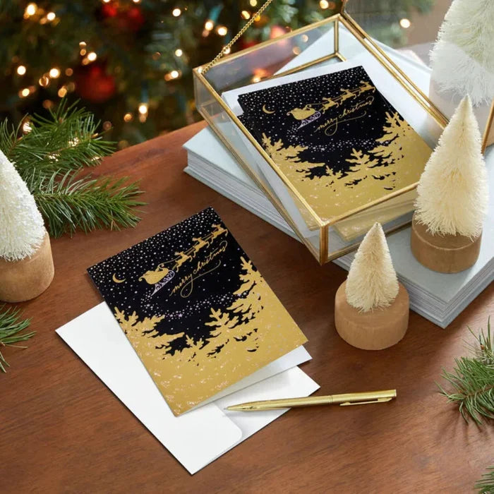 Decorative greeting cards with gold and black design on a wooden surface with Christmas decorations.