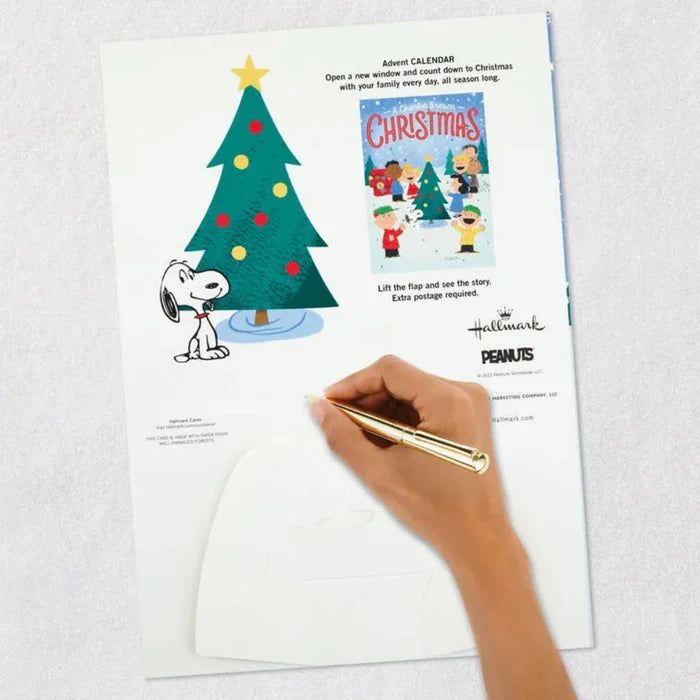 Hand holding a pen over a Christmas-themed card/advent calendar with a tree and cartoon character  from the peanuts gang on a white background.