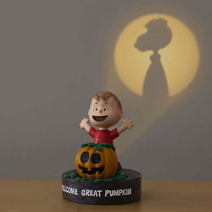 Figurine of a character in a red shirt with a pumpkin, standing on a base with 'Welcome Great Pumpkin' text, against a dark background with a shadow of a rooster.