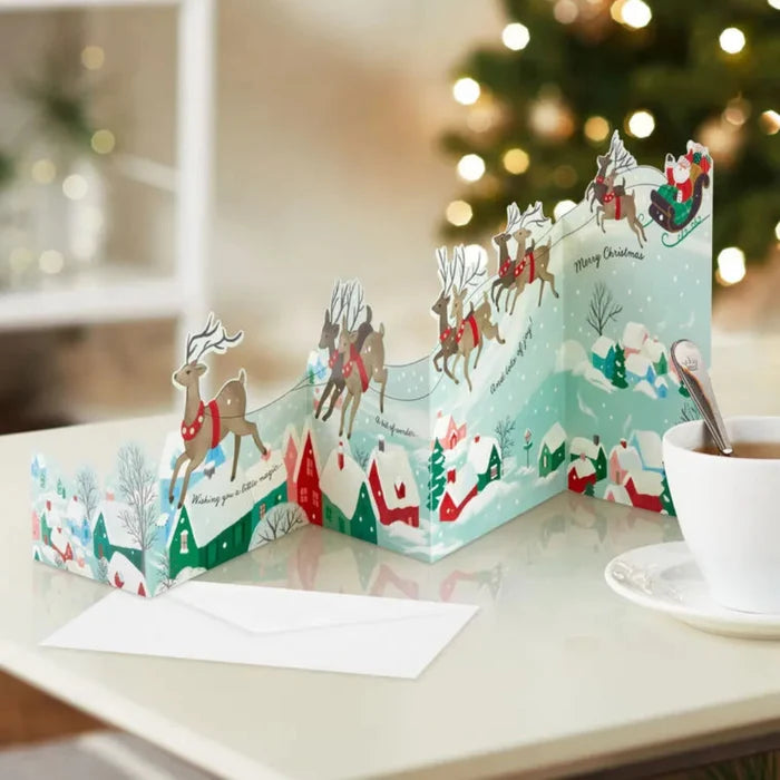 Christmas-themed pop-up card on a table with a cup of tea and saucer.
