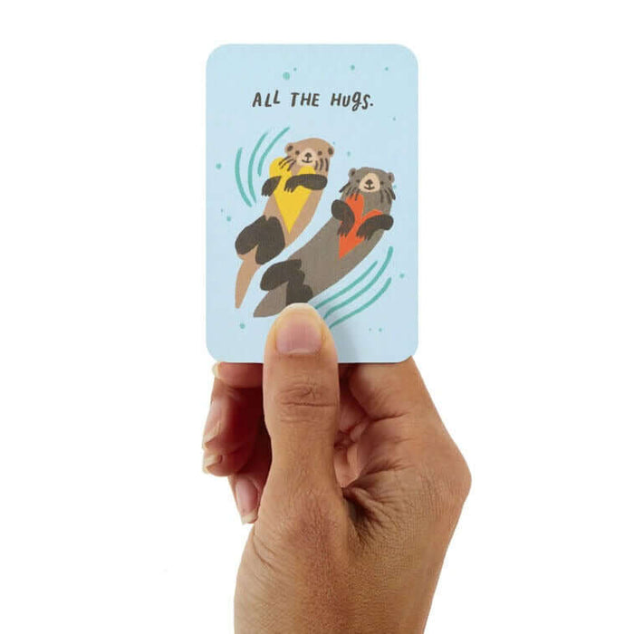 Greeting card with cartoon otters and text 'All the hugs' held by a hand on a white background