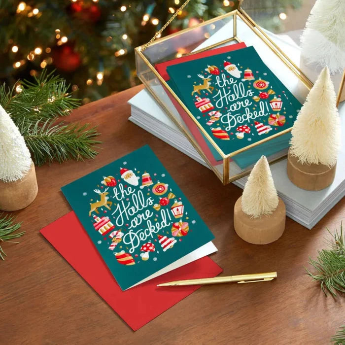 Christmas card with 'The Halls are Decked' design on a wooden surface with festive decorations.