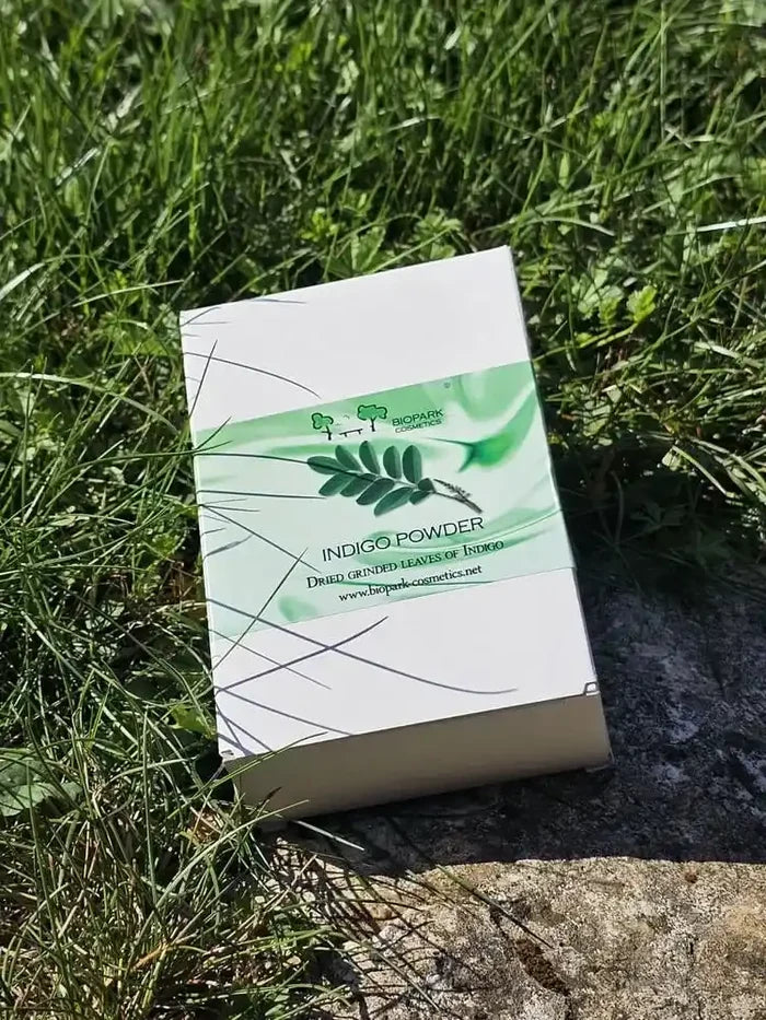 A white rectangular box of Indigo Powder (Dye Plant Leaves Extract) resting on a patch of green grass and stone outdoors.