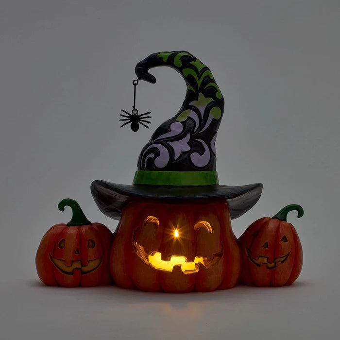 Pumpkin jack-o’-lantern glowing with LED light, wearing a decorative witch hat, with two smiling pumpkins on the sides.