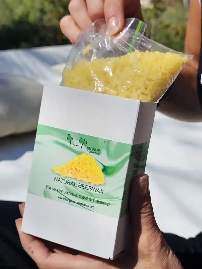 A person holds a white box labeled 'Natural Beeswax' with a clear plastic bag filled with yellow wax beads above the open box, photographed outdoors with a blurred light-colored surface and greenery in the background.