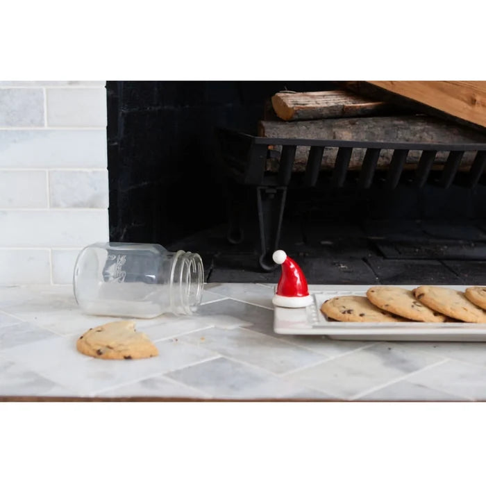 Cookies on a tray with a Santa hat next to an open jar in front of a fireplace.