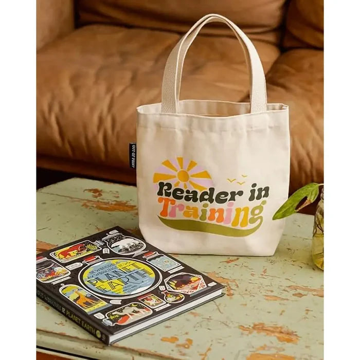 Tote bag with 'Reader in Training' text on a table next to a book