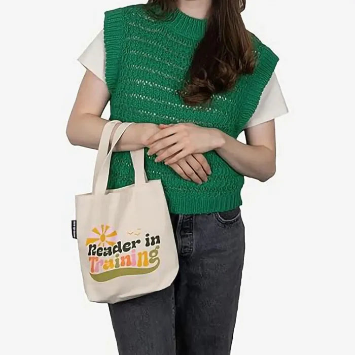 Person wearing a green knitted vest holding a tote bag with 'Reader in Training' text.