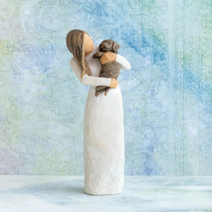 A Willow Tree style figurine of a woman in a long white dress holding a brown dog, set against a textured blue and white background.