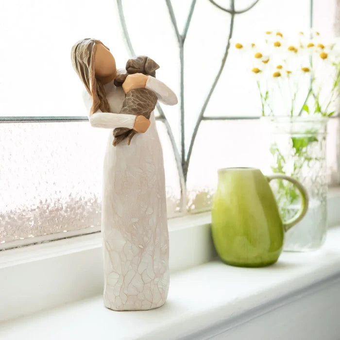 Decorative figurine of a woman holding a child on a windowsill with a green mug and flowers in the background.