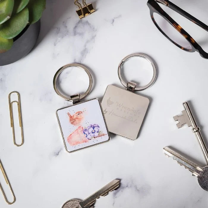 A square keychain displaying an artistic depiction of a bird among floral elements.