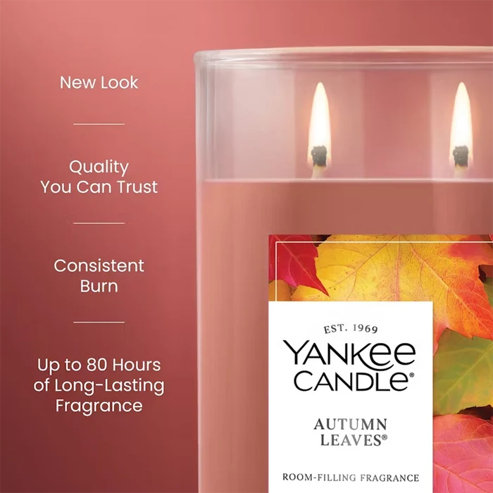 Close-up of the Yankee Candle Autumn Leaves tumbler with two flames lit. On the left side of the image, text highlights features: “New Look, Quality You Can Trust, Consistent Burn, Up to 80 Hours of Long-Lasting Fragrance.”
